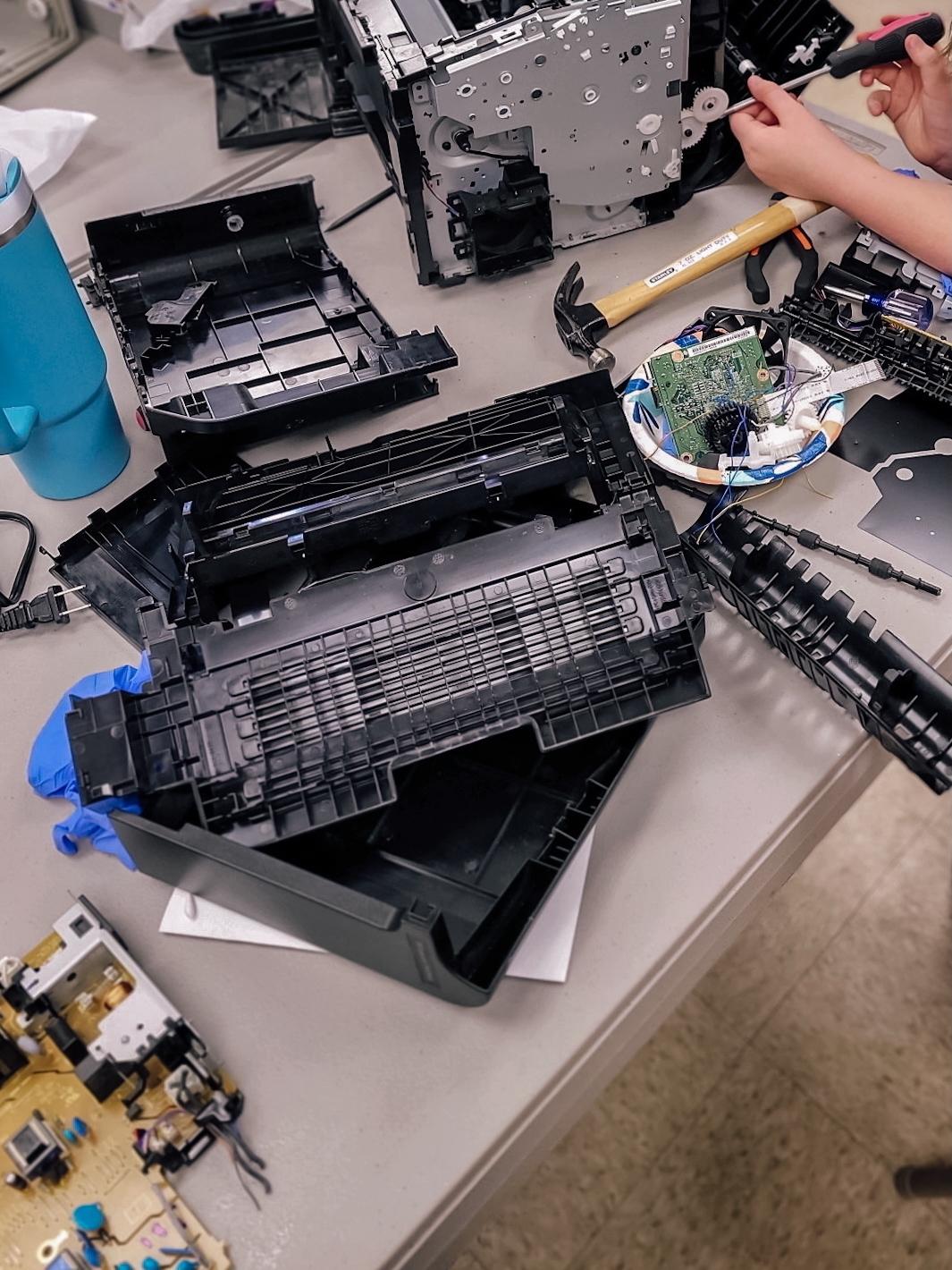 Auto-generated description: A person is disassembling a printer on a table cluttered with various parts and tools.