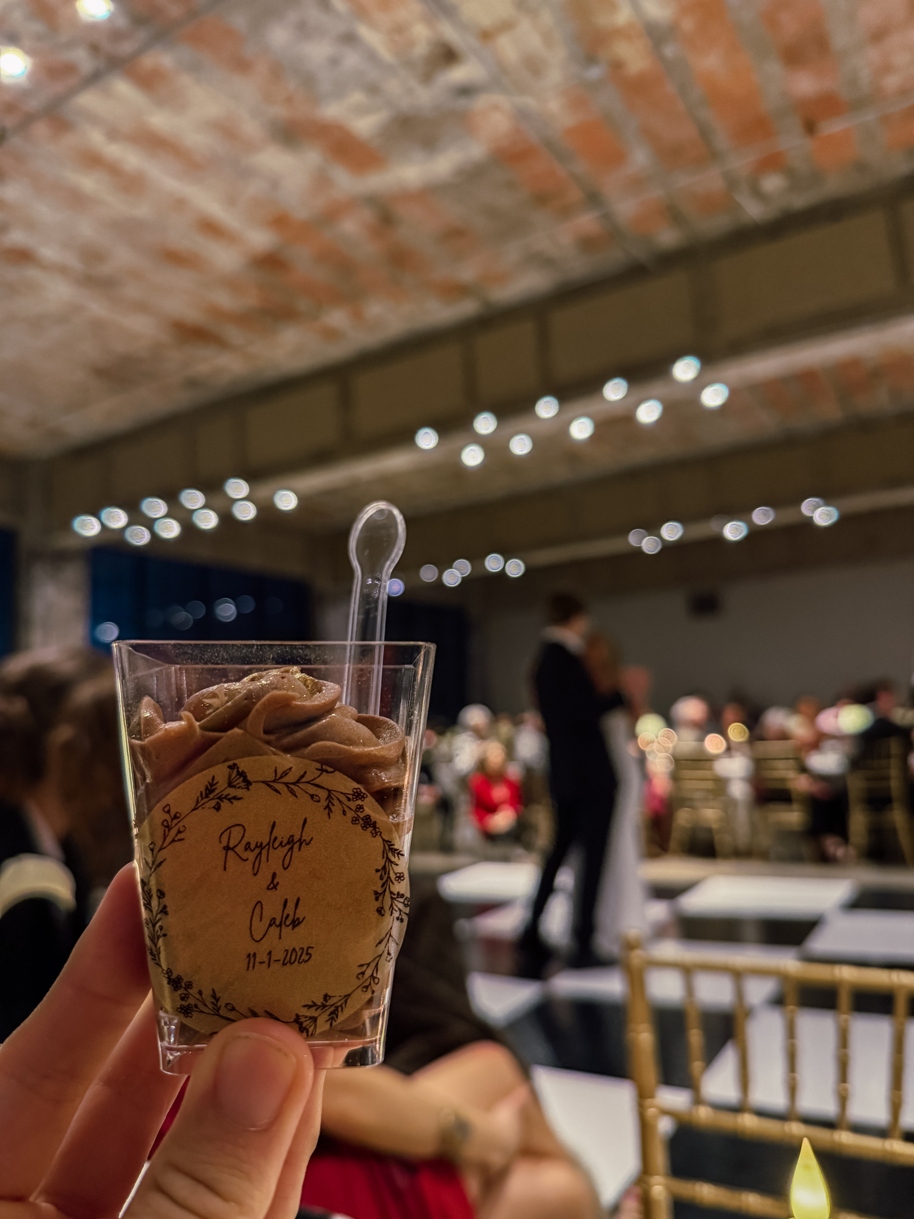 A chocolate dessert in a clear cup is held up, with a couple dancing in the blurred background of a decorated reception hall.