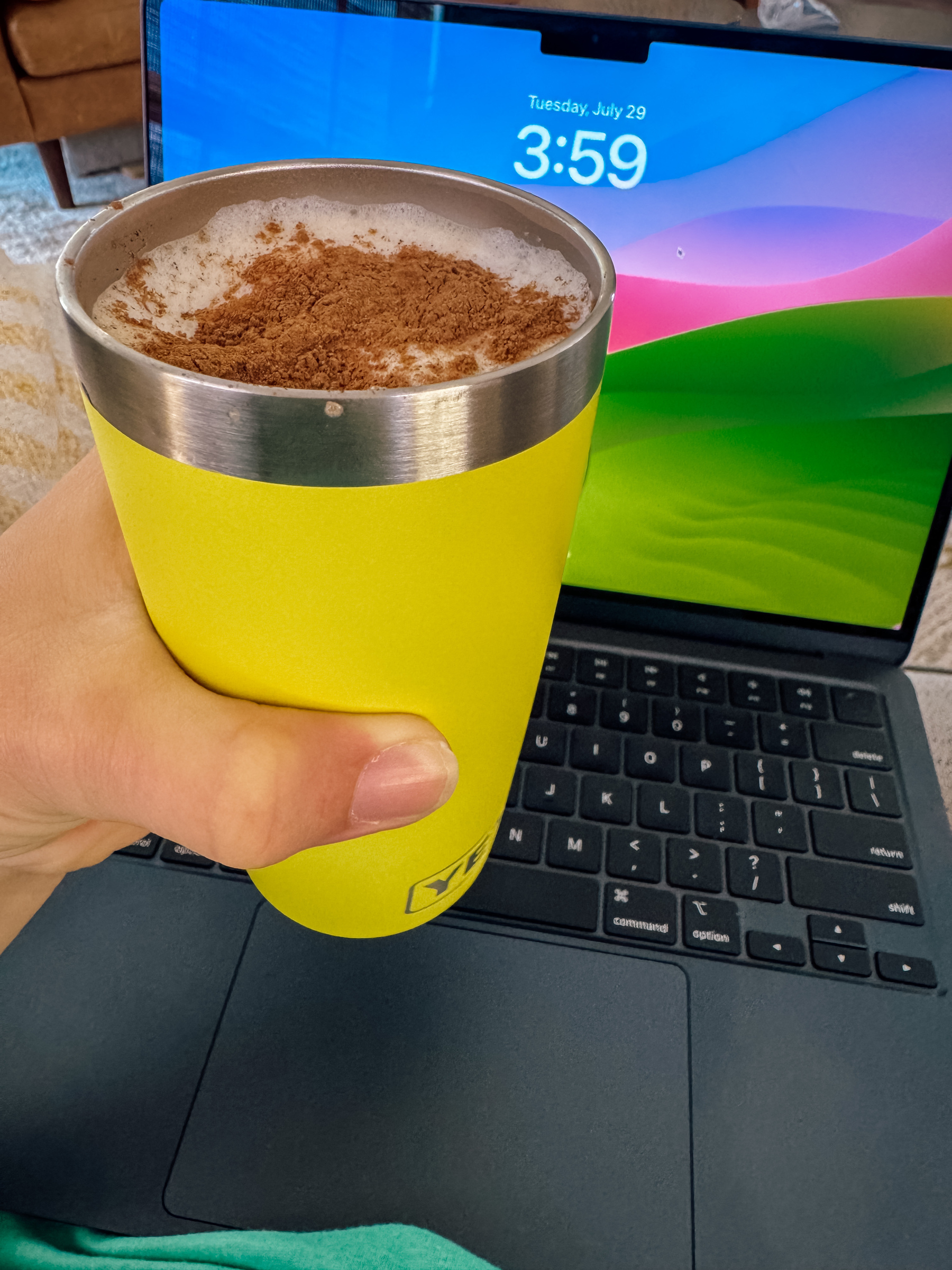 A hand holds a brightly colored cup filled with a drink topped with frothy milk and cinnamon with a brightly colored laptop screen in the background