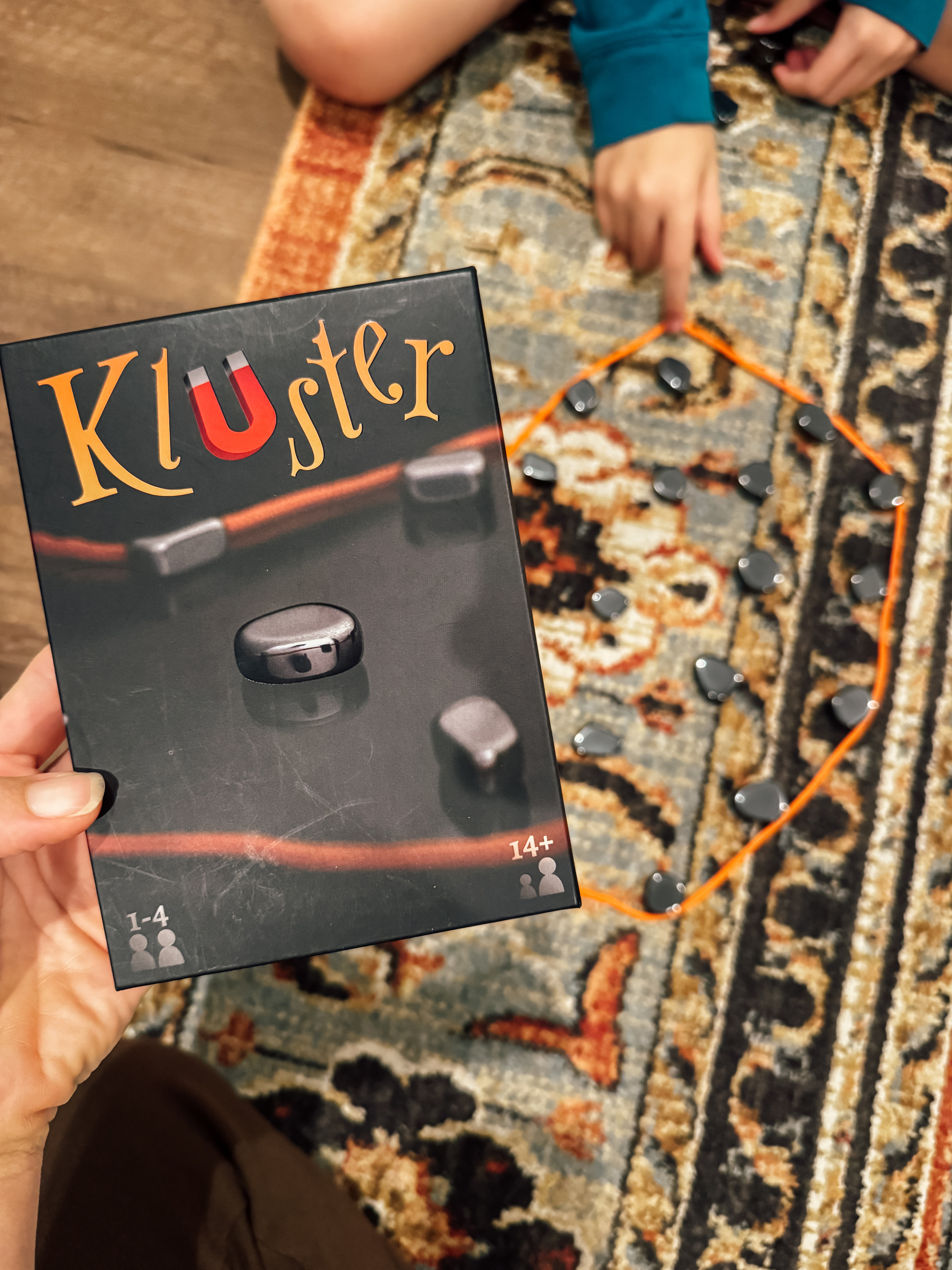 A person holds the box of a game called "Kluster" while the game pieces are arranged in a circle on a patterned carpet.