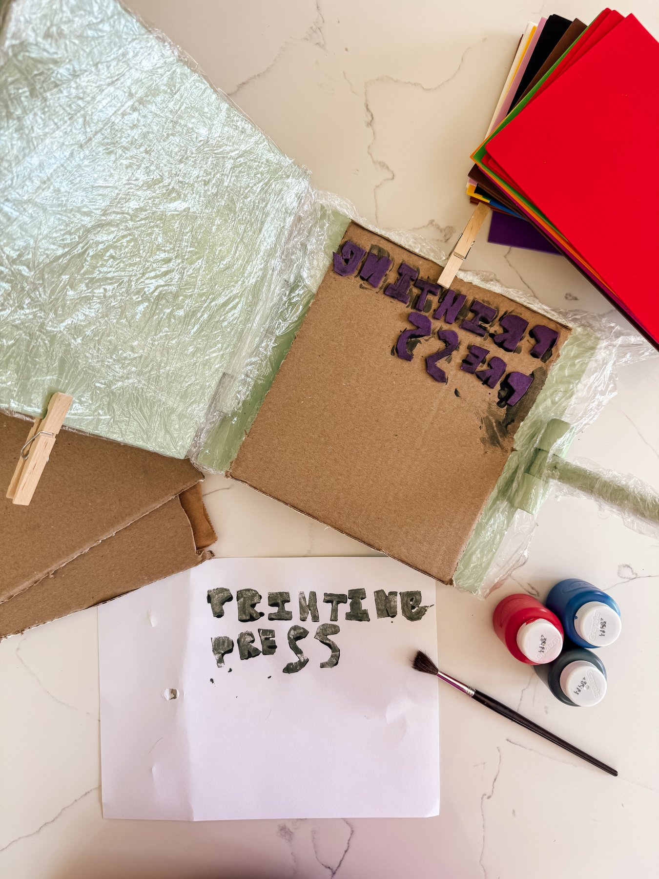 Auto-generated description: A handmade printing press setup is shown with painted text on paper, cardboard, paintbrush, and small paint containers.