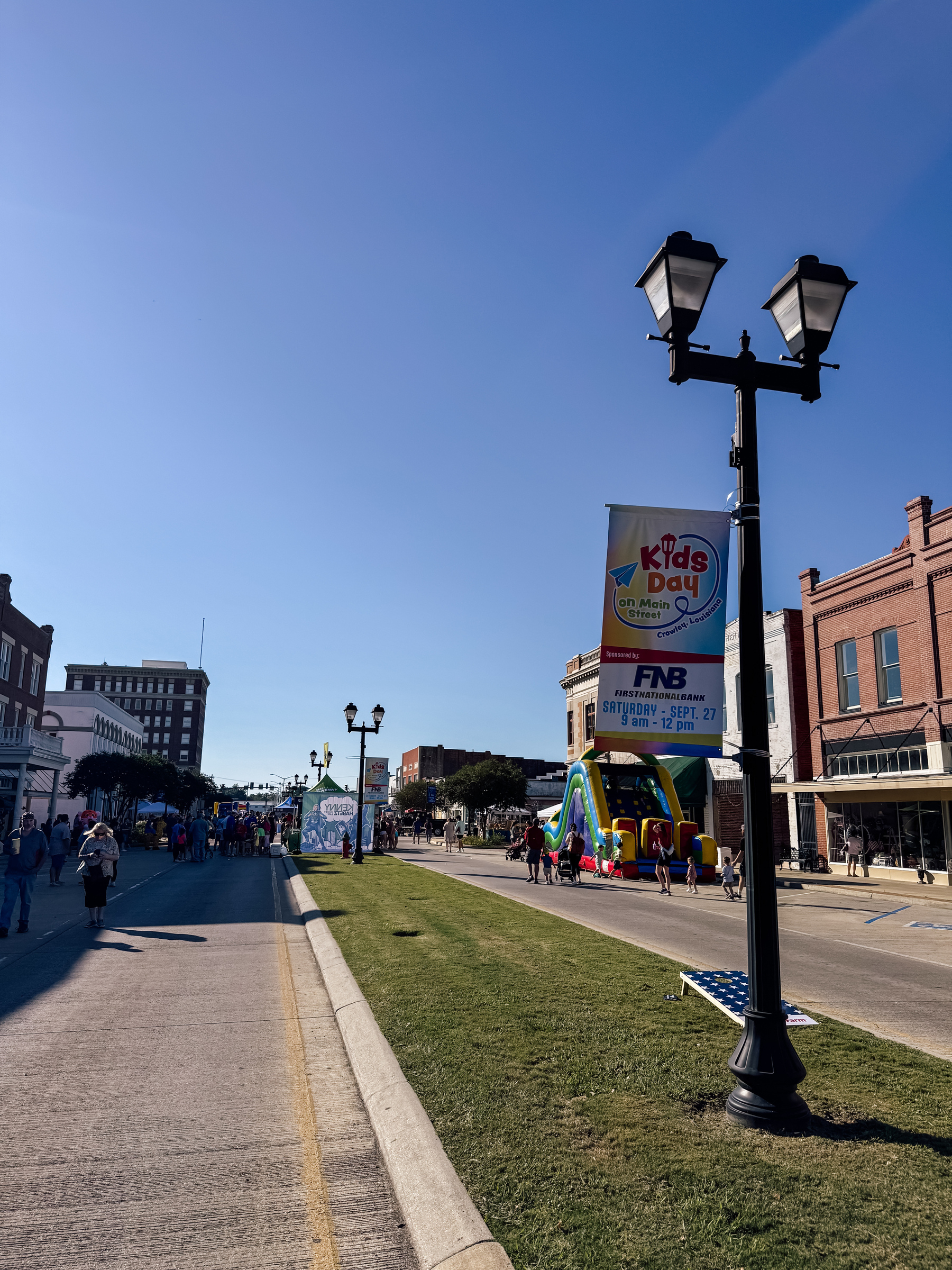 A street scene featuring a festival with a Kids Day banner, inflatable play structures, and people walking along a road.