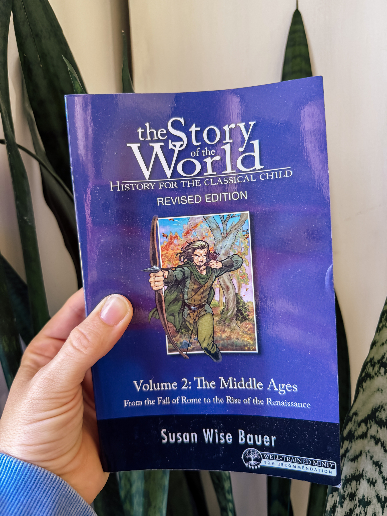 Auto-generated description: A hand is holding the book The Story of the World: History for the Classical Child, Volume 2: The Middle Ages by Susan Wise Bauer, with a blue cover featuring an illustration of an archer.