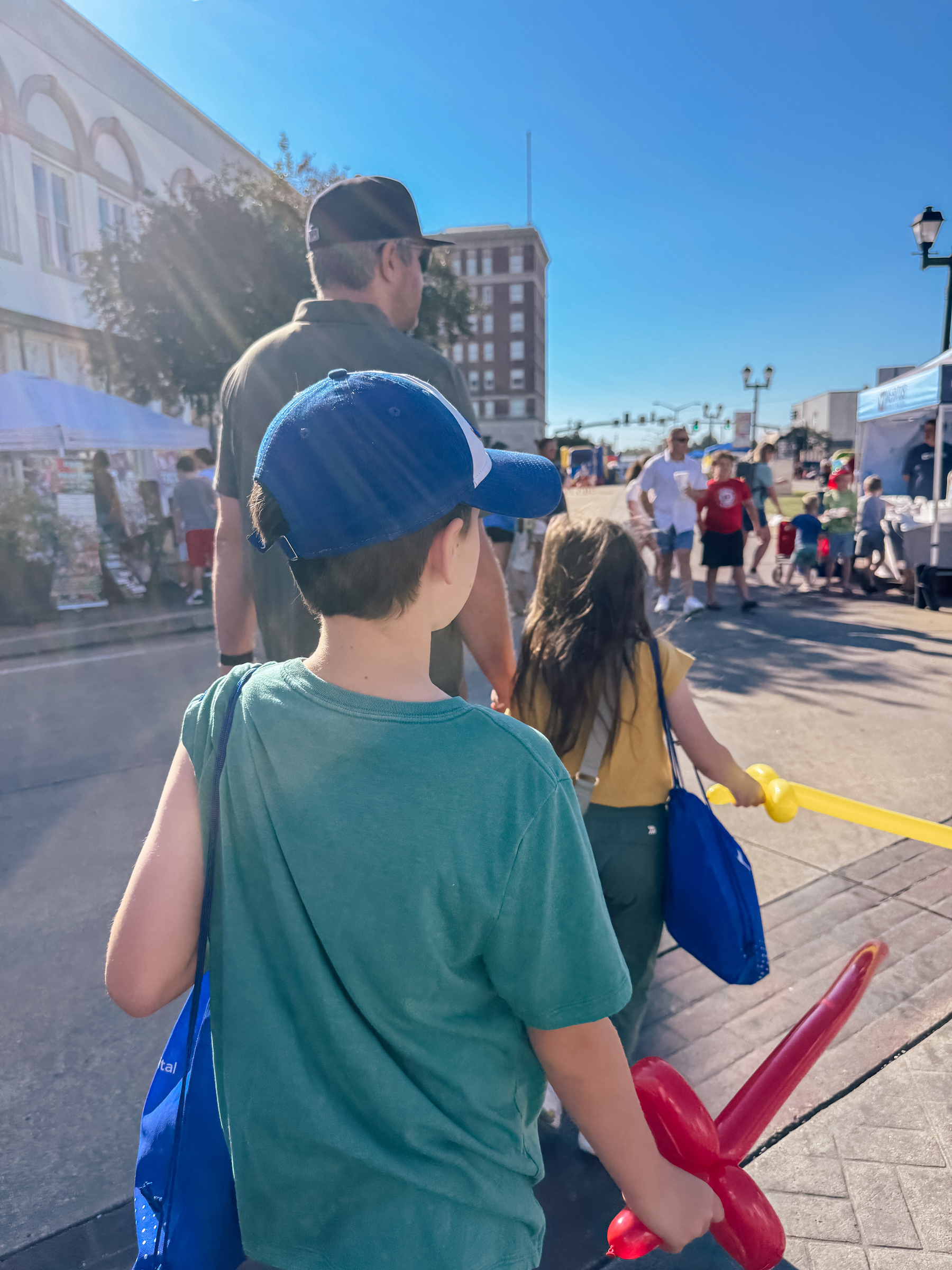 A group of people, including a man and children with balloon animals, walk through an outdoor market or event on a sunny day.