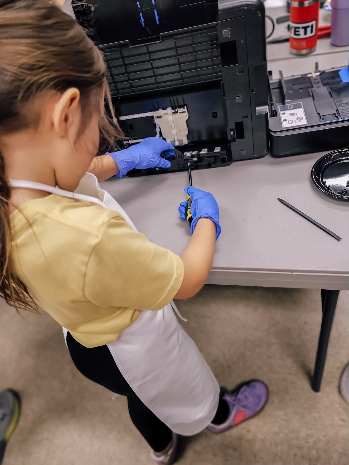 Auto-generated description: A child wearing gloves and an apron is disassembling a piece of equipment on a table.