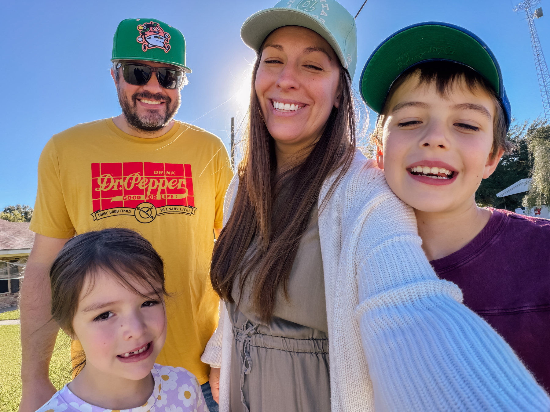 A family of four, with two adults and two children, smiles while posing for a selfie outdoors on a sunny day.