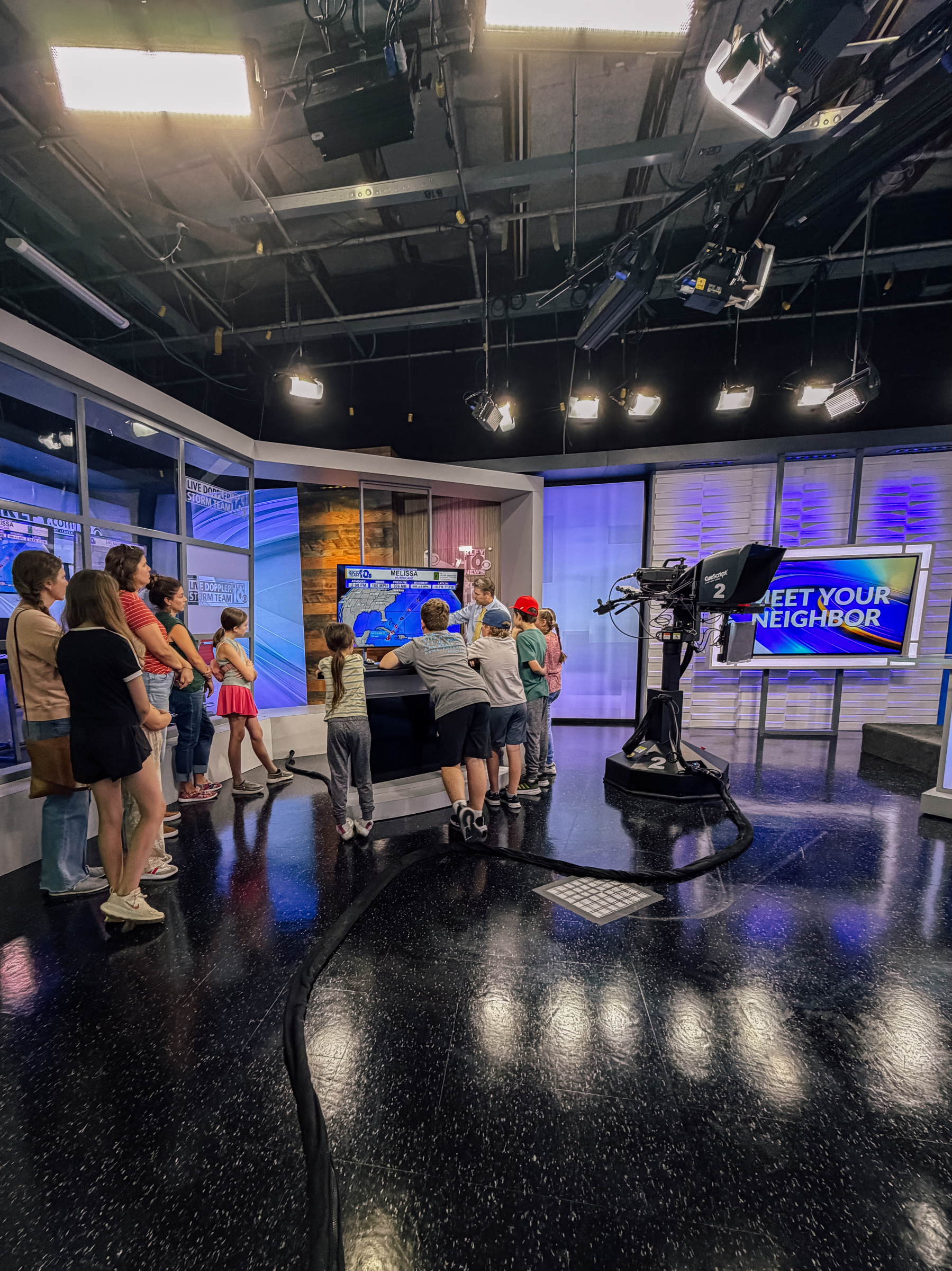 A group of children and adults gather around a weather station setup in a studio with bright lighting and large screens.