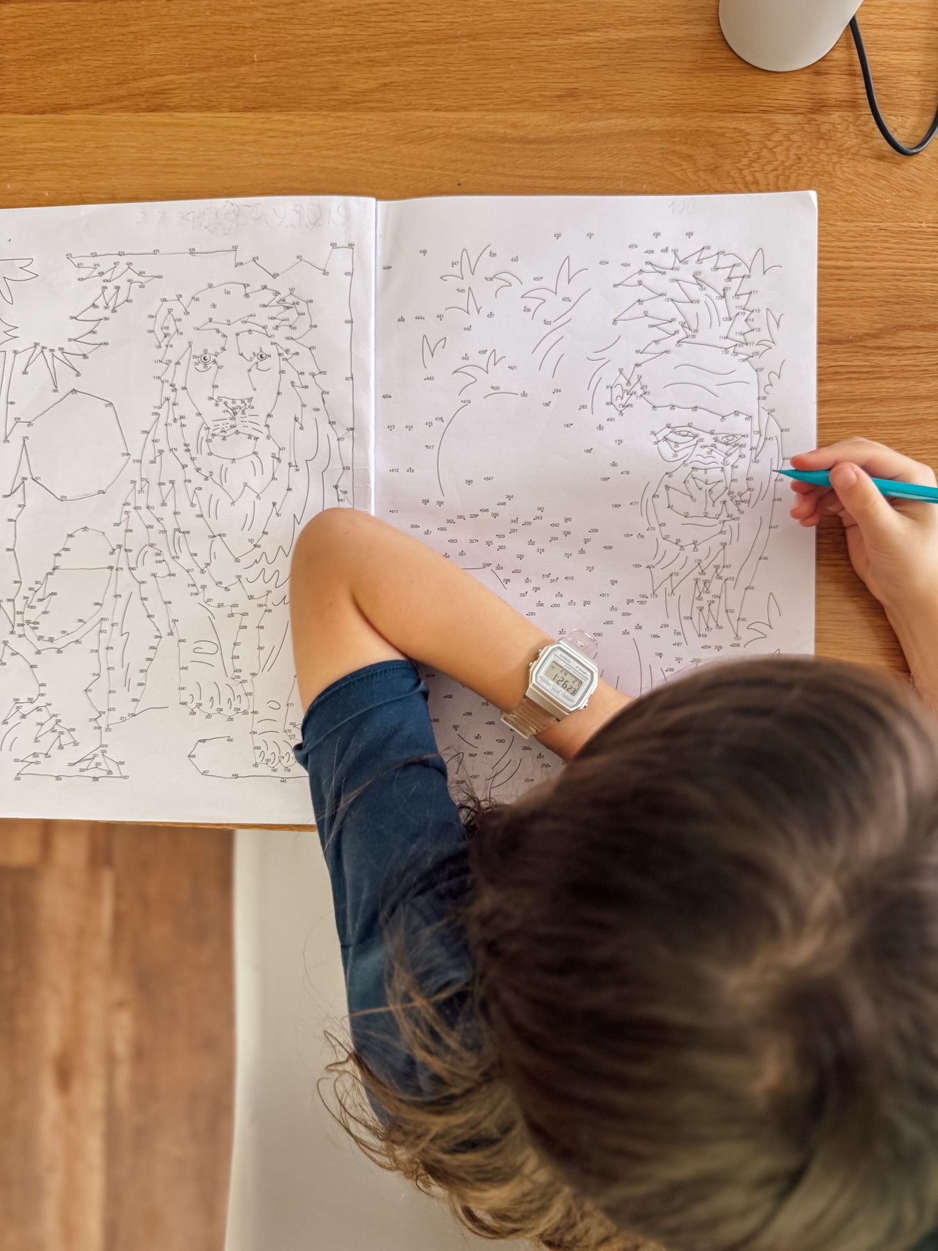 Auto-generated description: A child is drawing in a dot-to-dot coloring book at a wooden table.
