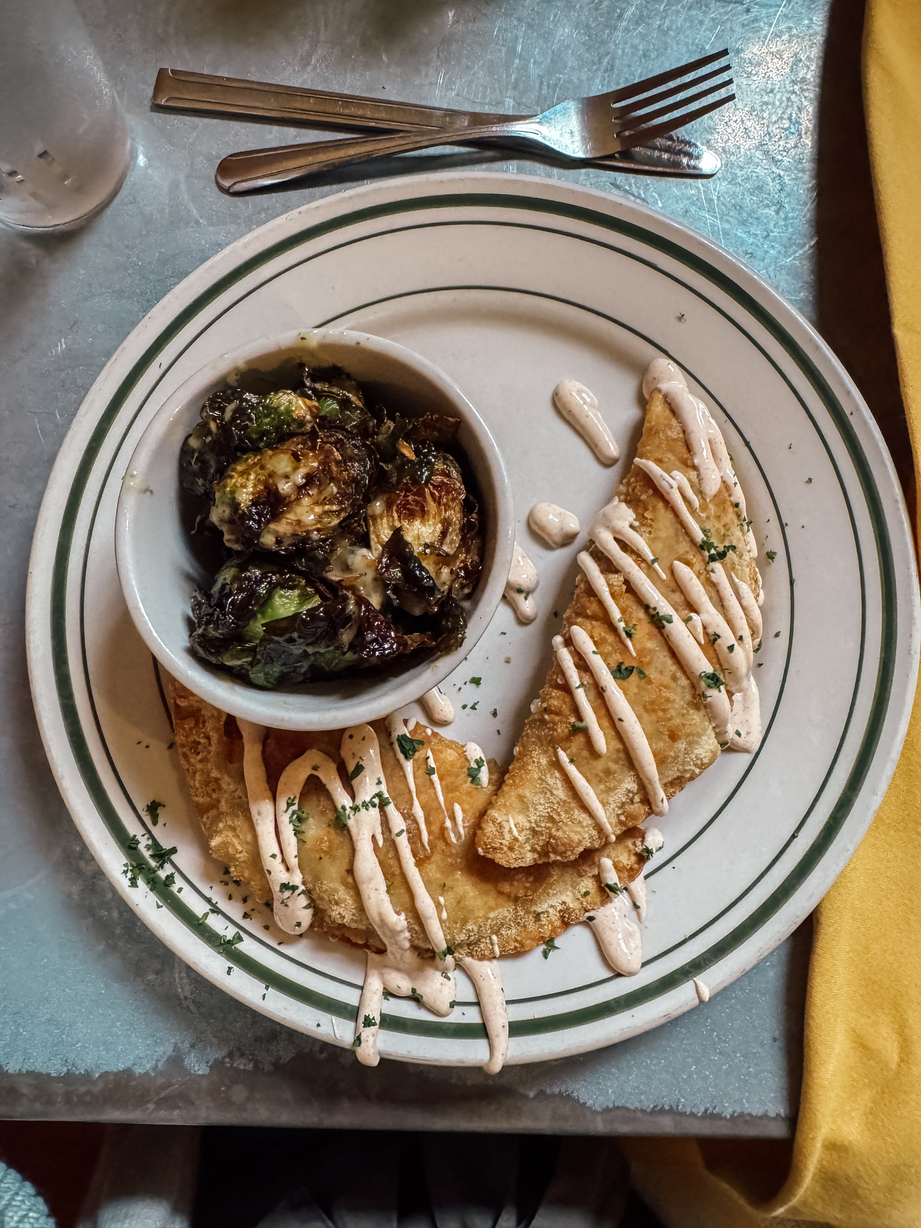 A plate of hand pies drizzled with sauce is served alongside a small bowl of roasted Brussels sprouts.