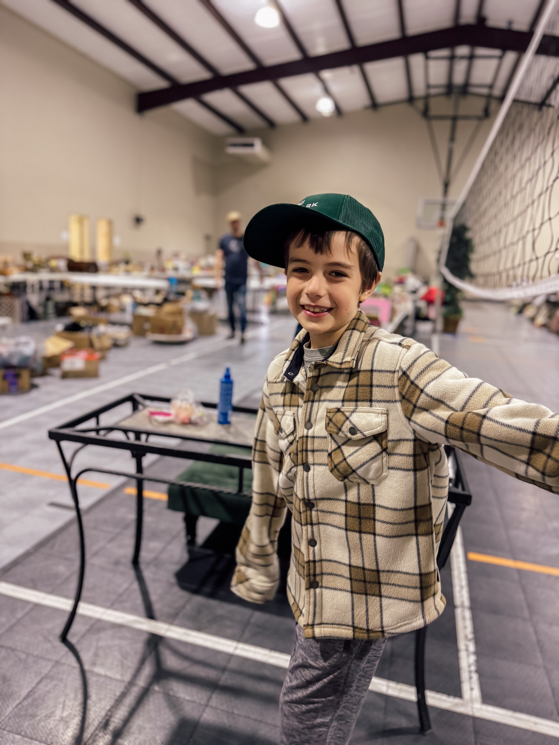 A child wearing a plaid jacket and green cap is smiling in a large indoor space with tables and various items in the background.