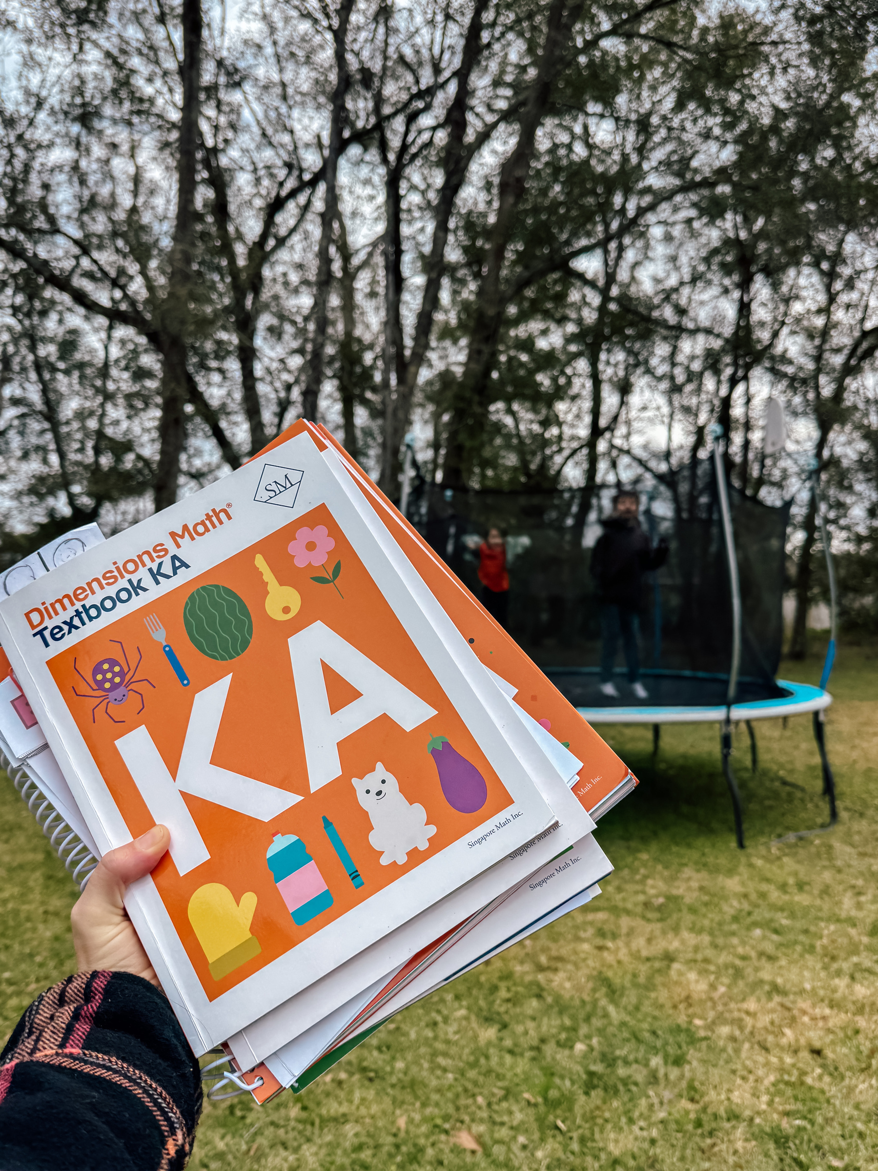 A person is holding school textbooks outdoors near a trampoline with children playing on it.