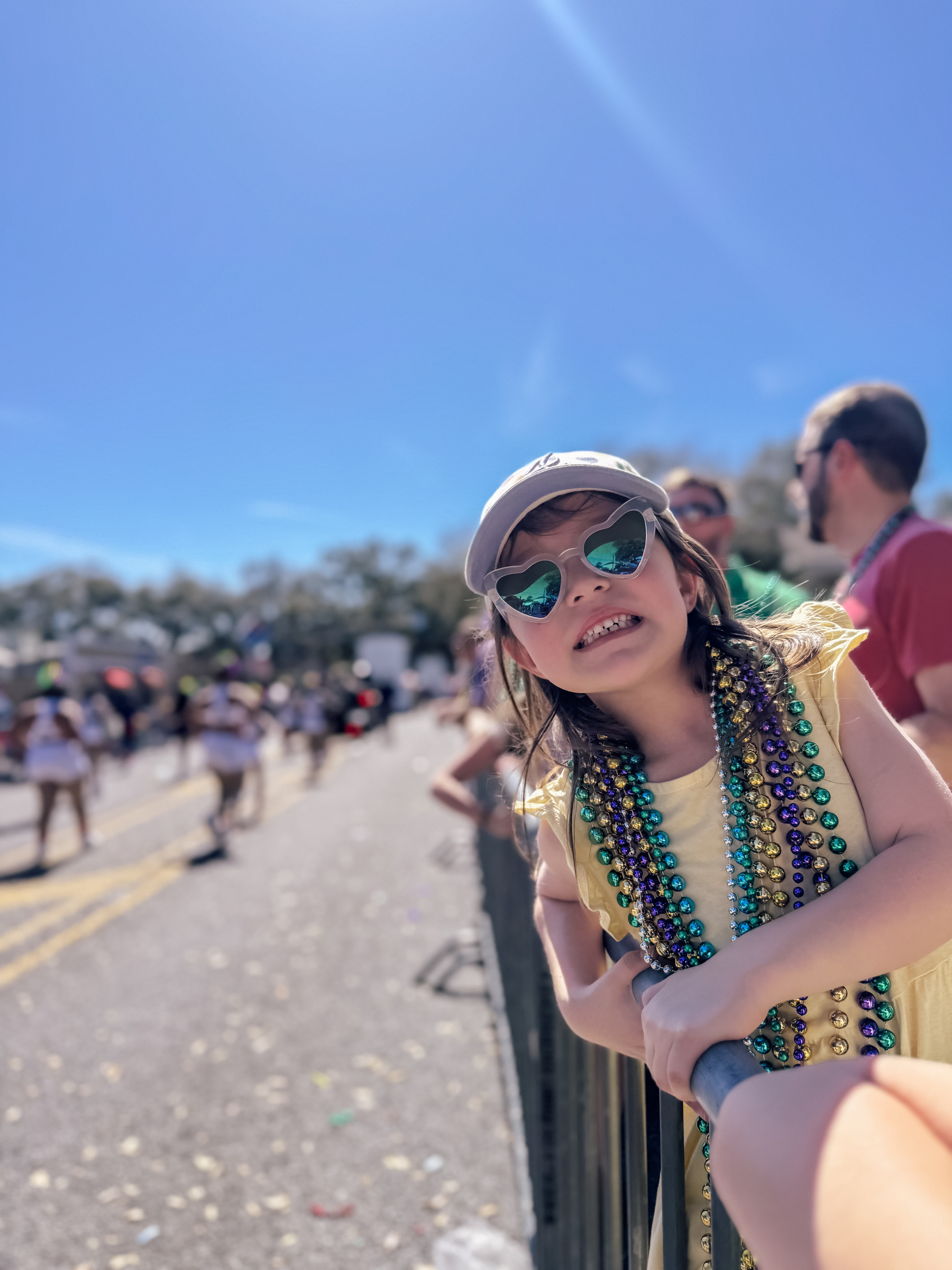 A young child wearing a cap, heart-shaped sunglasses, and colorful beads smiles while leaning against a railing during a Mardi Gras parade. 