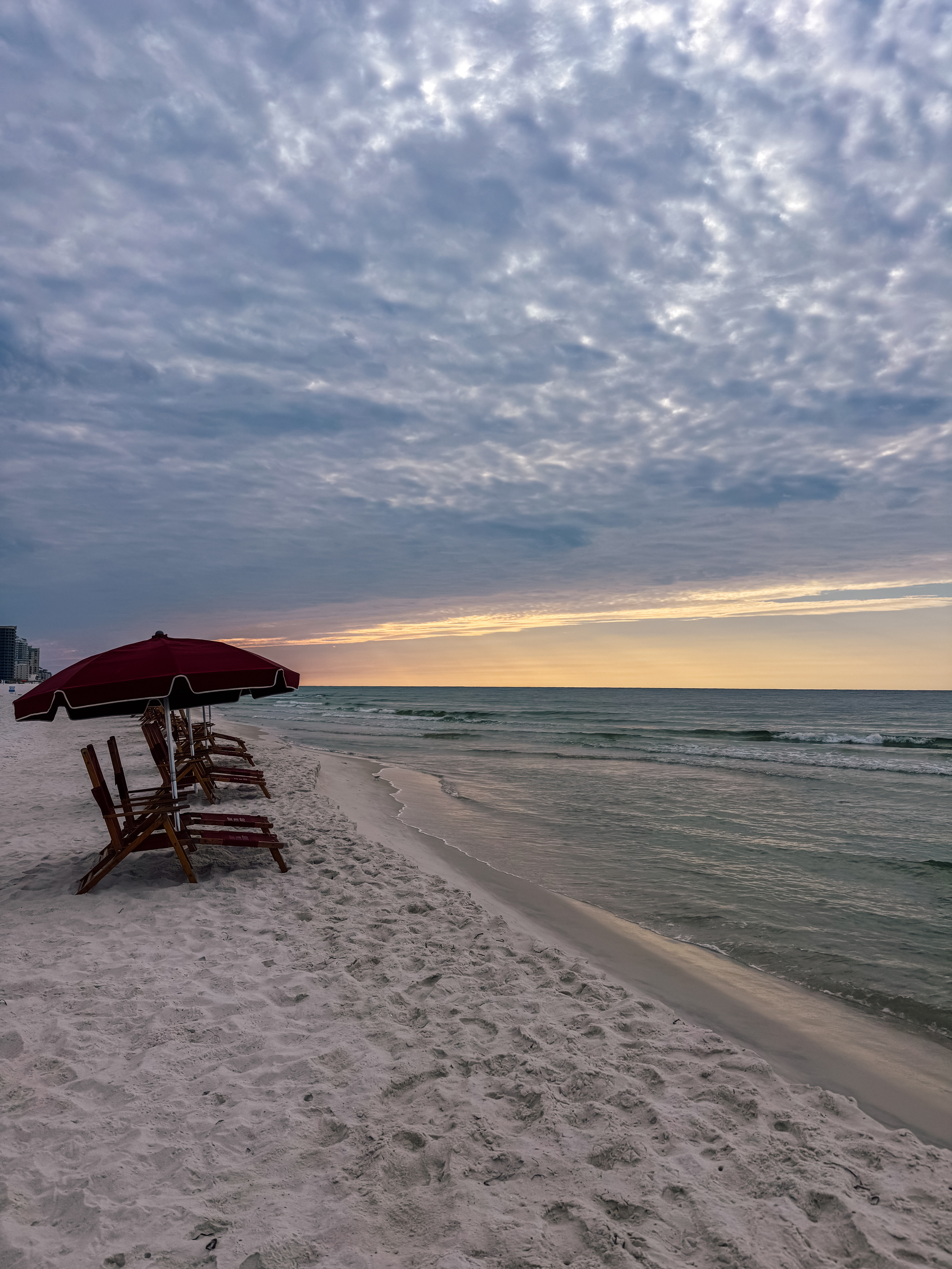 A line of wooden beach chairs with red umbrellas faces the ocean on a sandy beach under a dramatic, cloudy sky at sunrise.