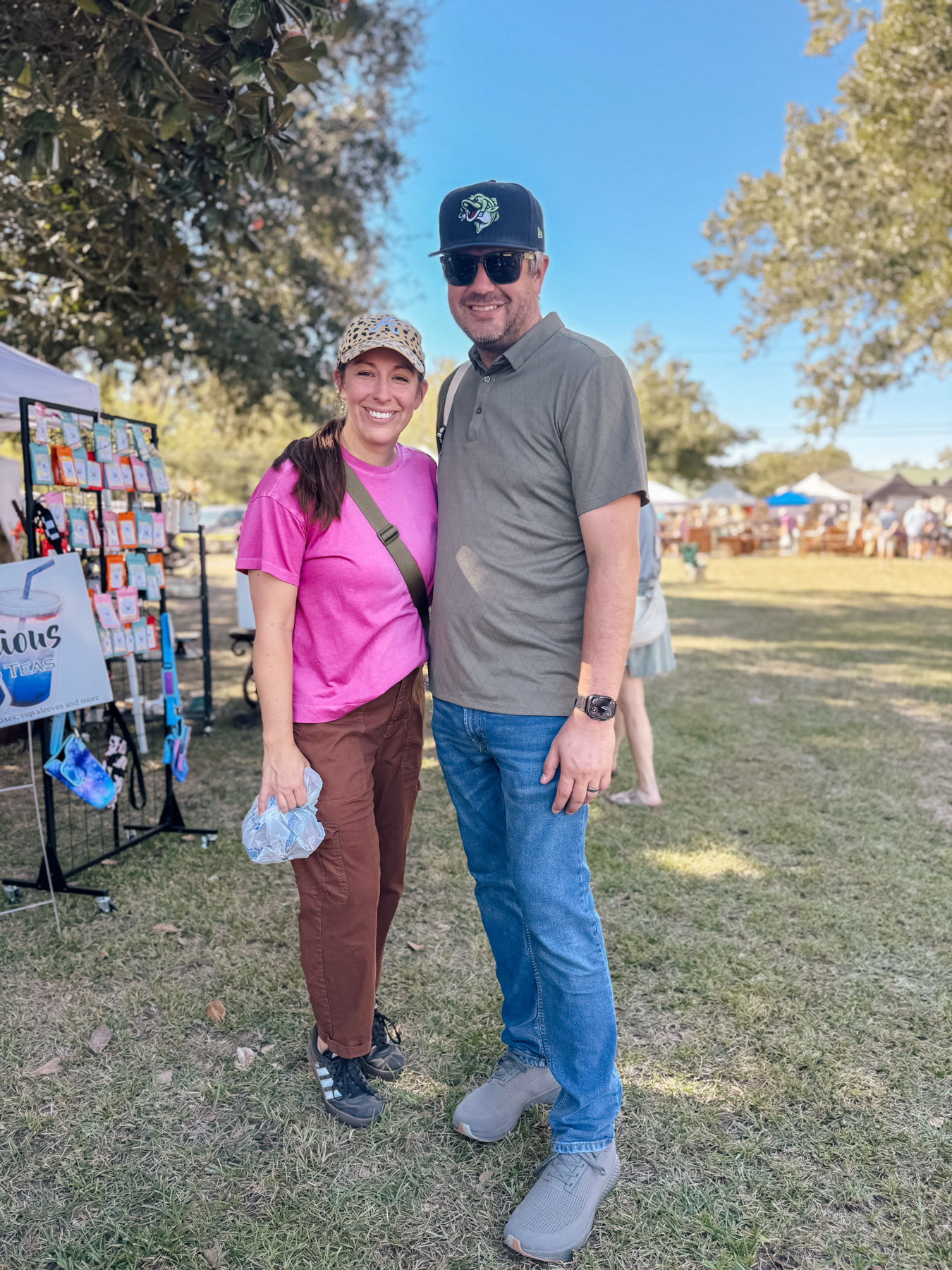 Auto-generated description: A smiling couple is posing together outdoors at a park event with booths in the background.