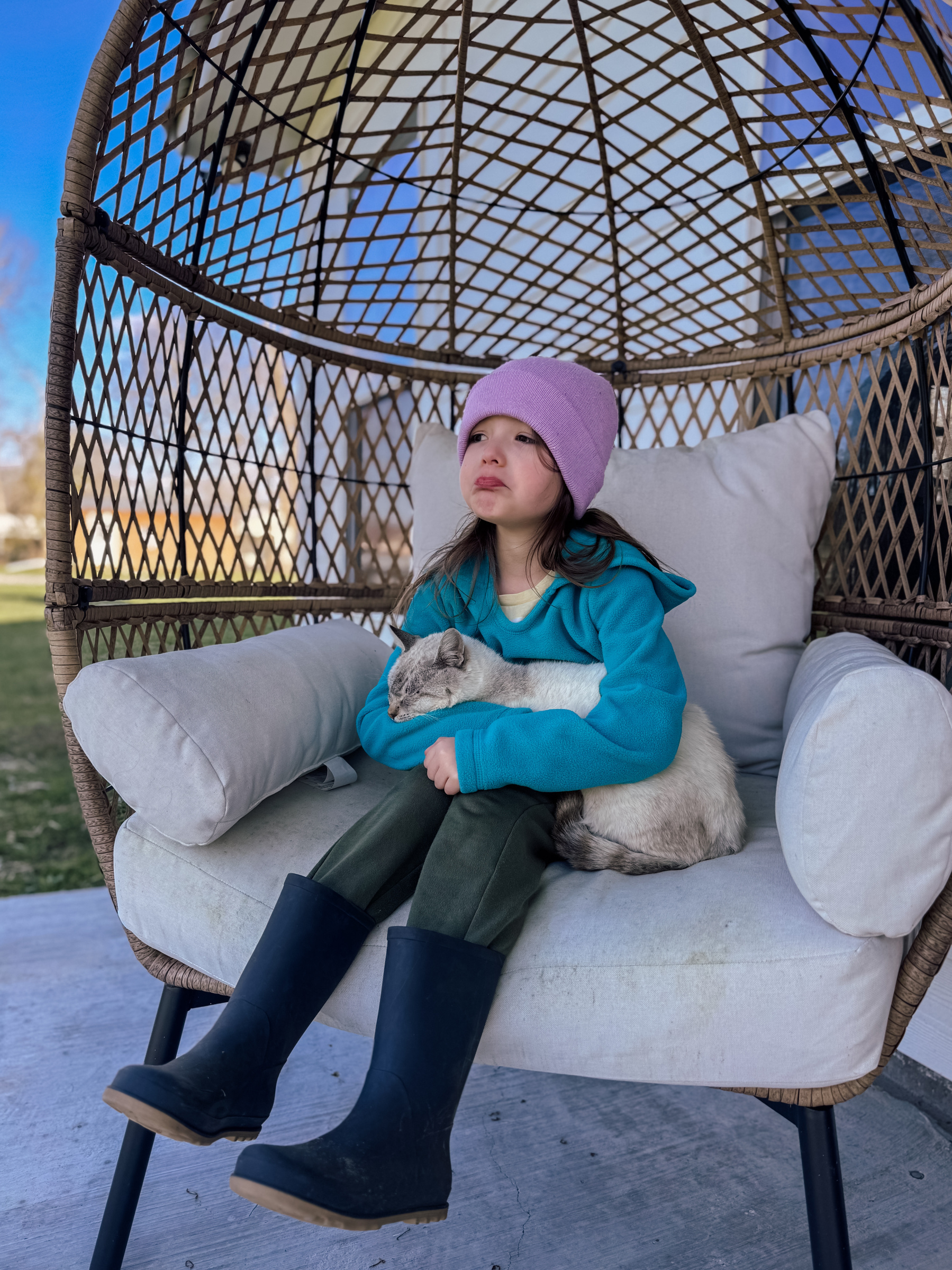 A child wearing a purple beanie and teal jacket sits on a cushioned outdoor chair, holding a cat.