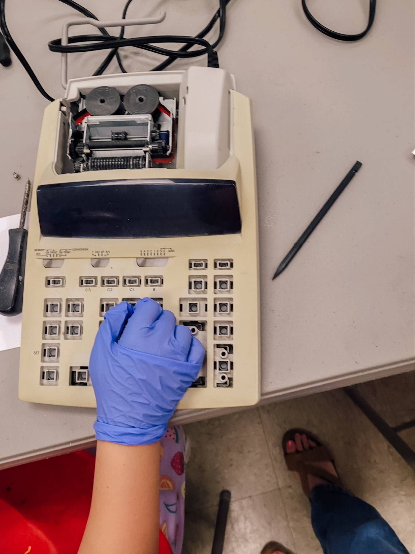 Auto-generated description: A person wearing a blue glove is disassembling a vintage printing calculator with an open top, surrounded by tools and wires.