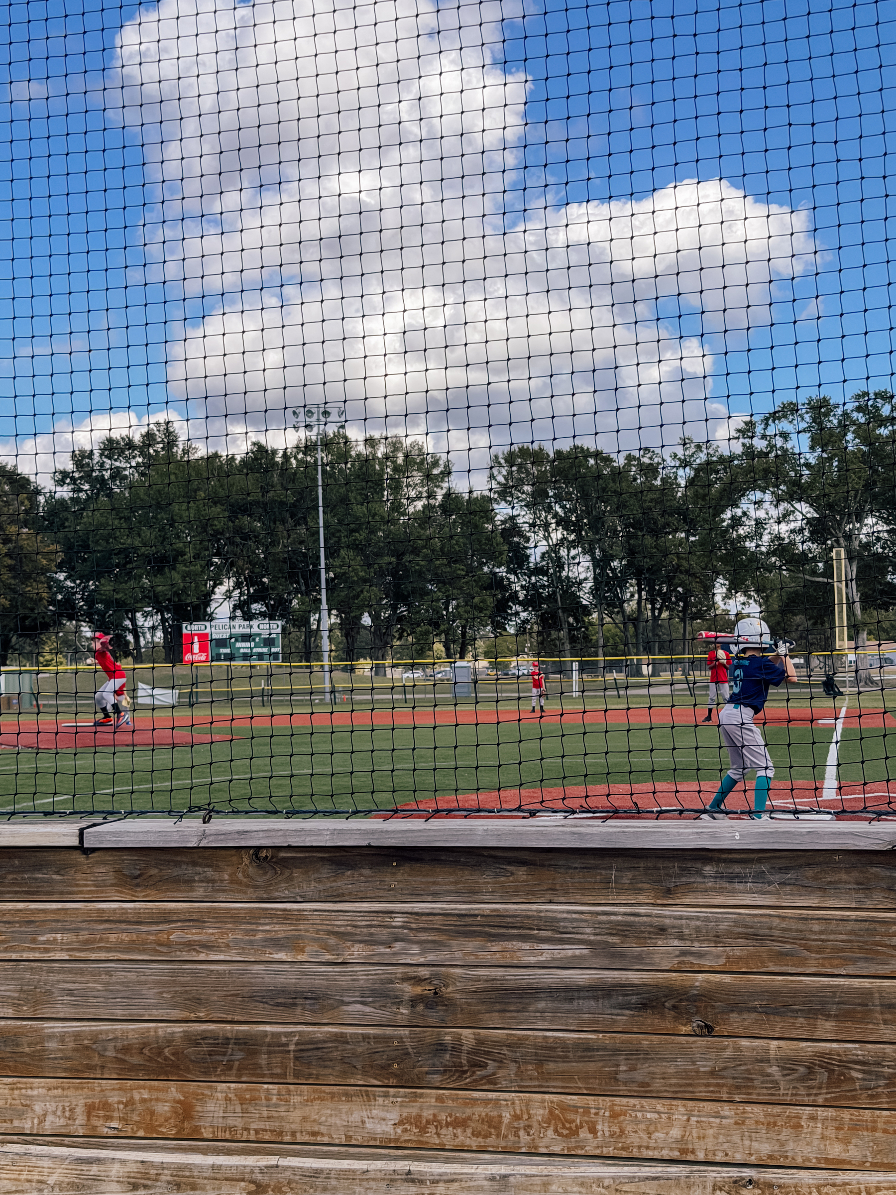 A youth baseball game is taking place on a sunny day, viewed through a protective netting.