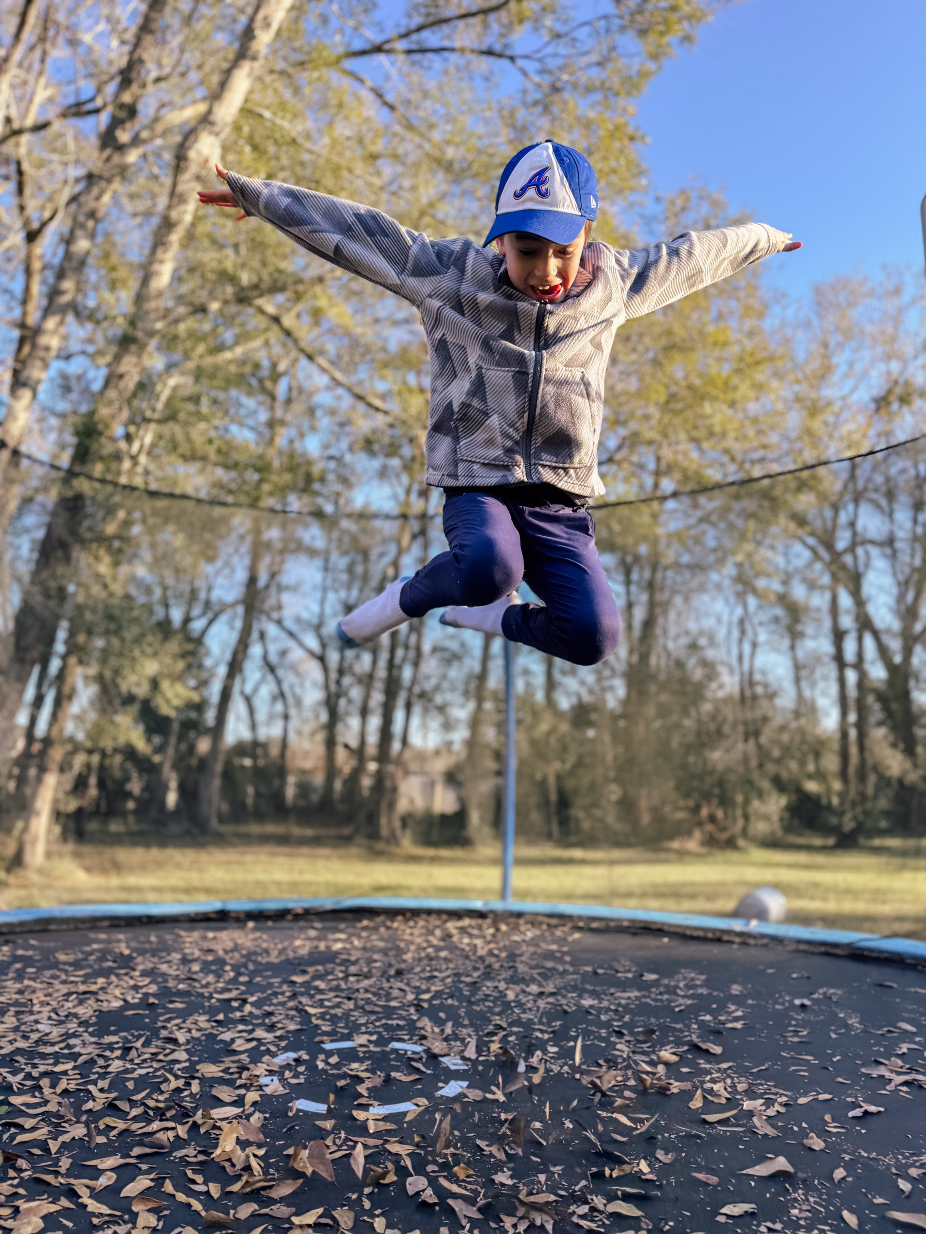 A child wearing a cap and jacket joyfully jumps on a leaf-covered trampoline surrounded by trees.