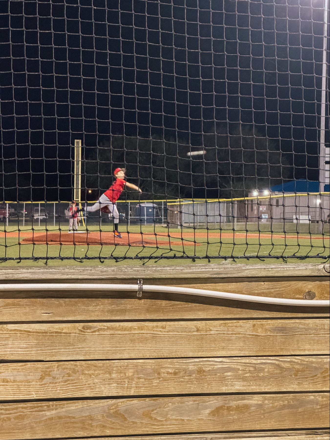 Auto-generated description: A baseball player in a red uniform is pitching the ball on a field at night, viewed through a net.