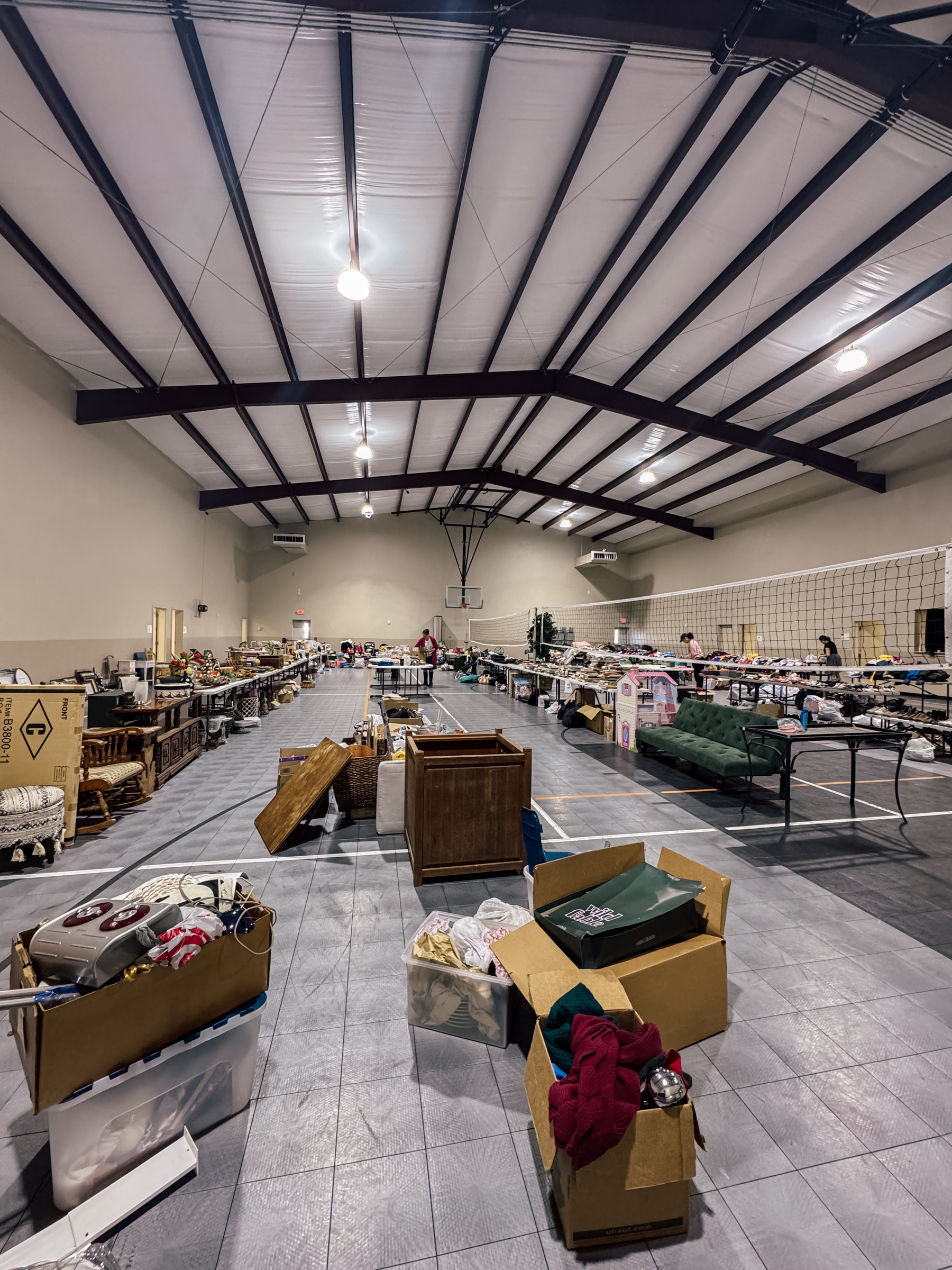 A large indoor space is filled with tables displaying various items for sale, resembling a garage sale or flea market.