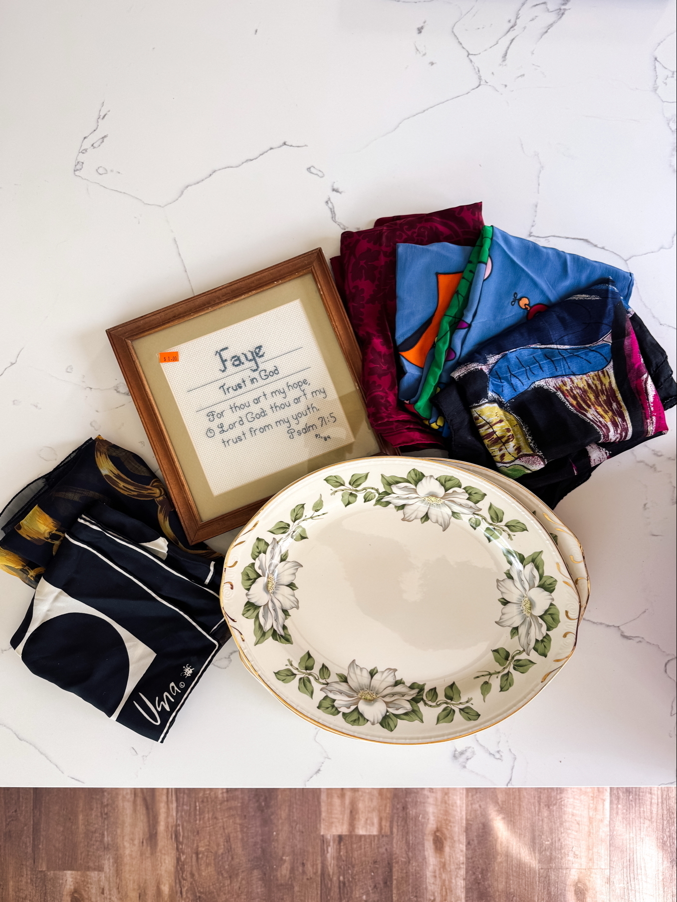 Auto-generated description: A floral-patterned platter is placed beside a stack of colorful folded scarves and a framed handwritten note.