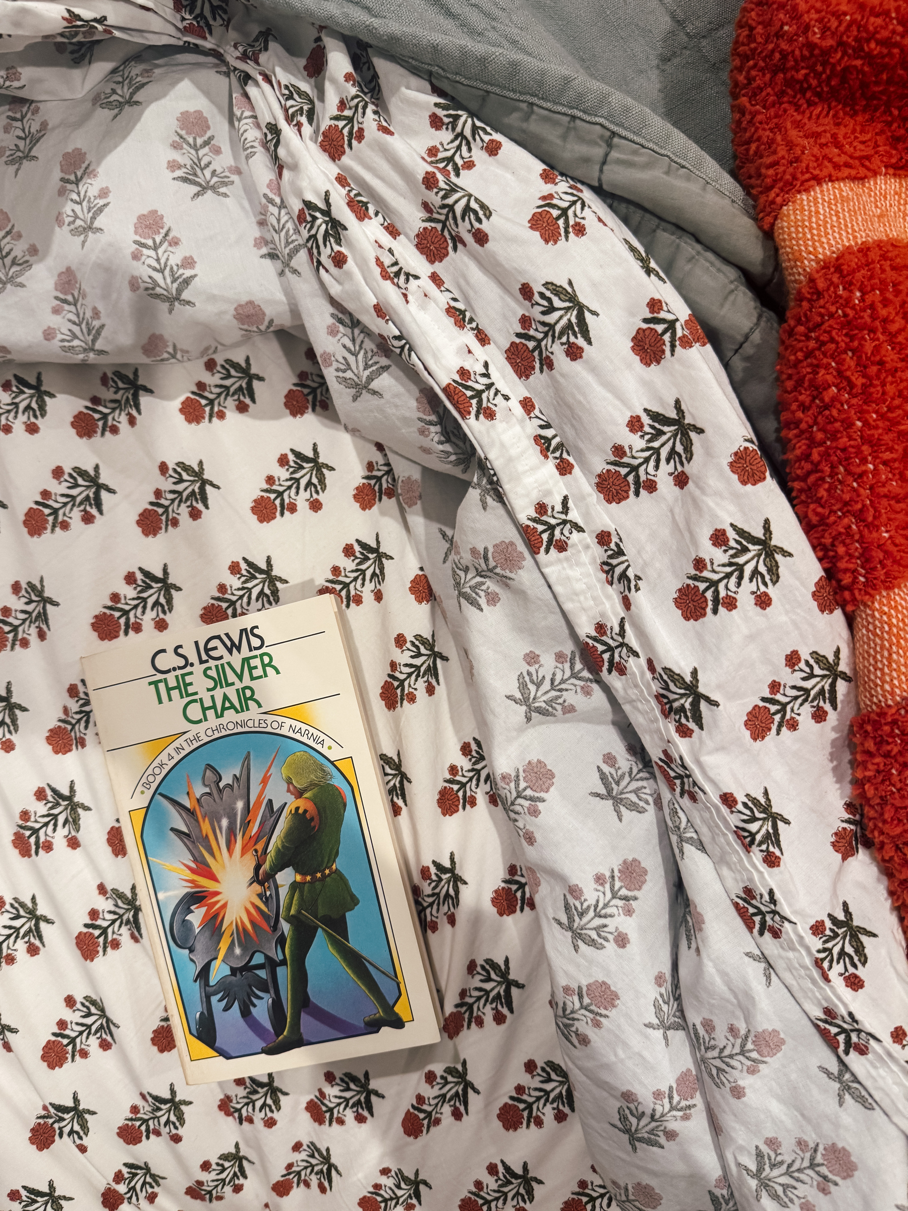 A book titled "The Silver Chair" by C.S. Lewis rests on a floral-patterned bedsheet next to a red-orange textured blanket.