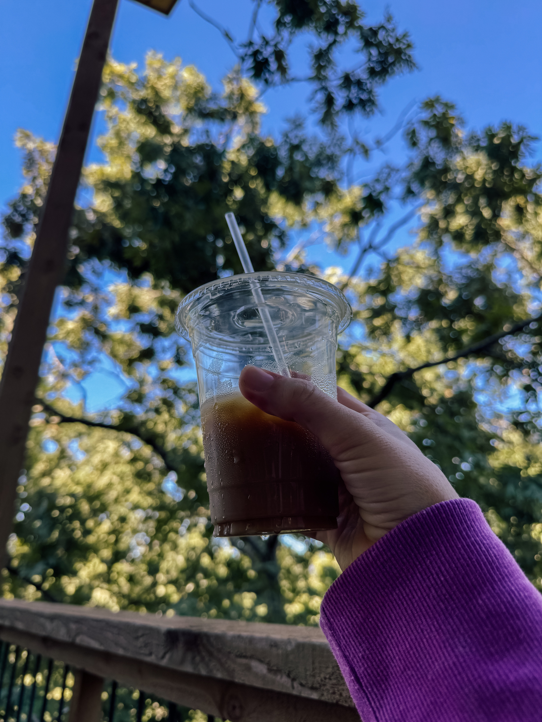 A hand in a purple sleeve holds a plastic cup with a straw against a backdrop of green trees and a clear blue sky.