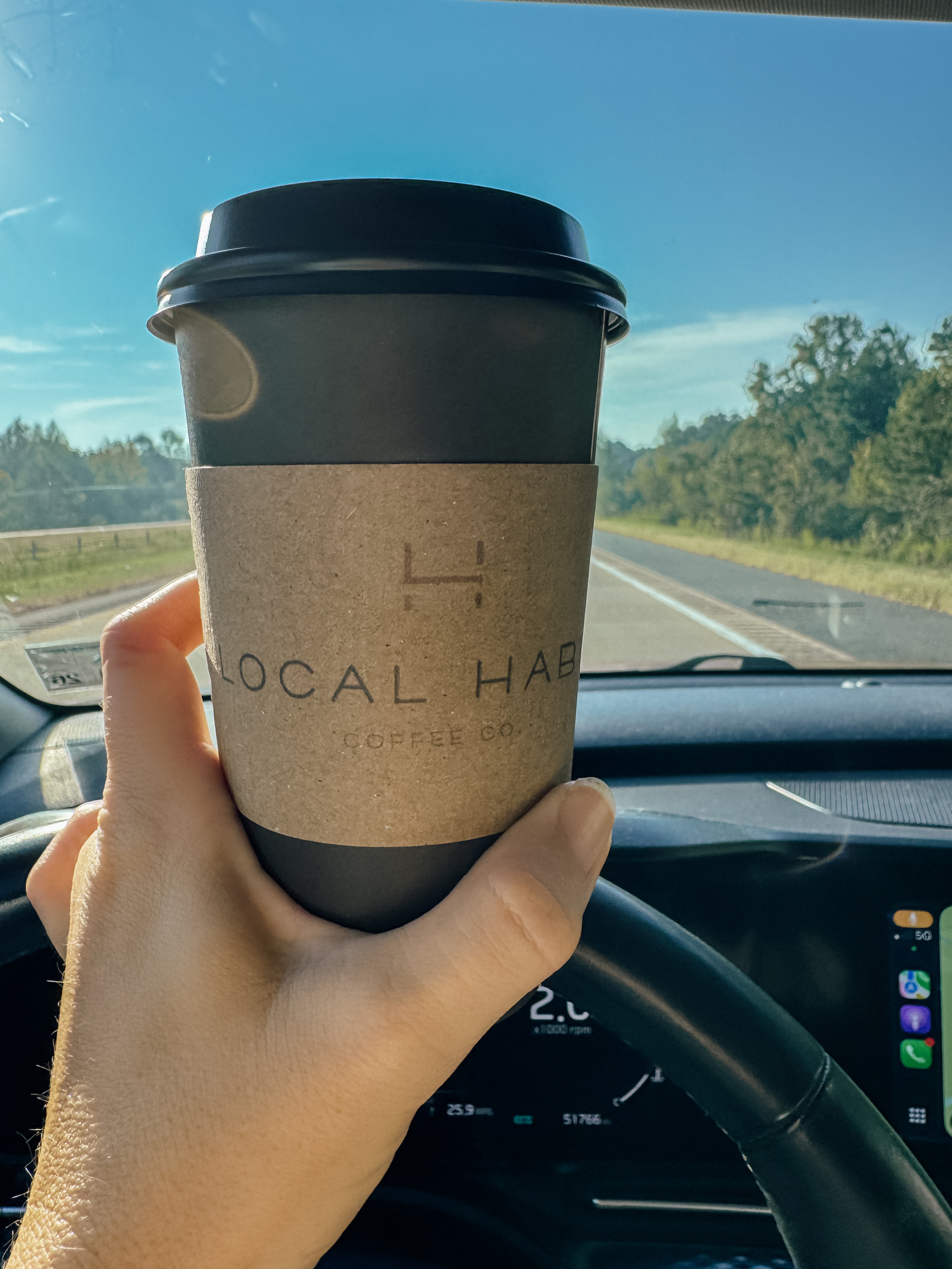 A hand is holding a coffee cup labeled "Local Habit" while driving on a sunny road with trees in the background.