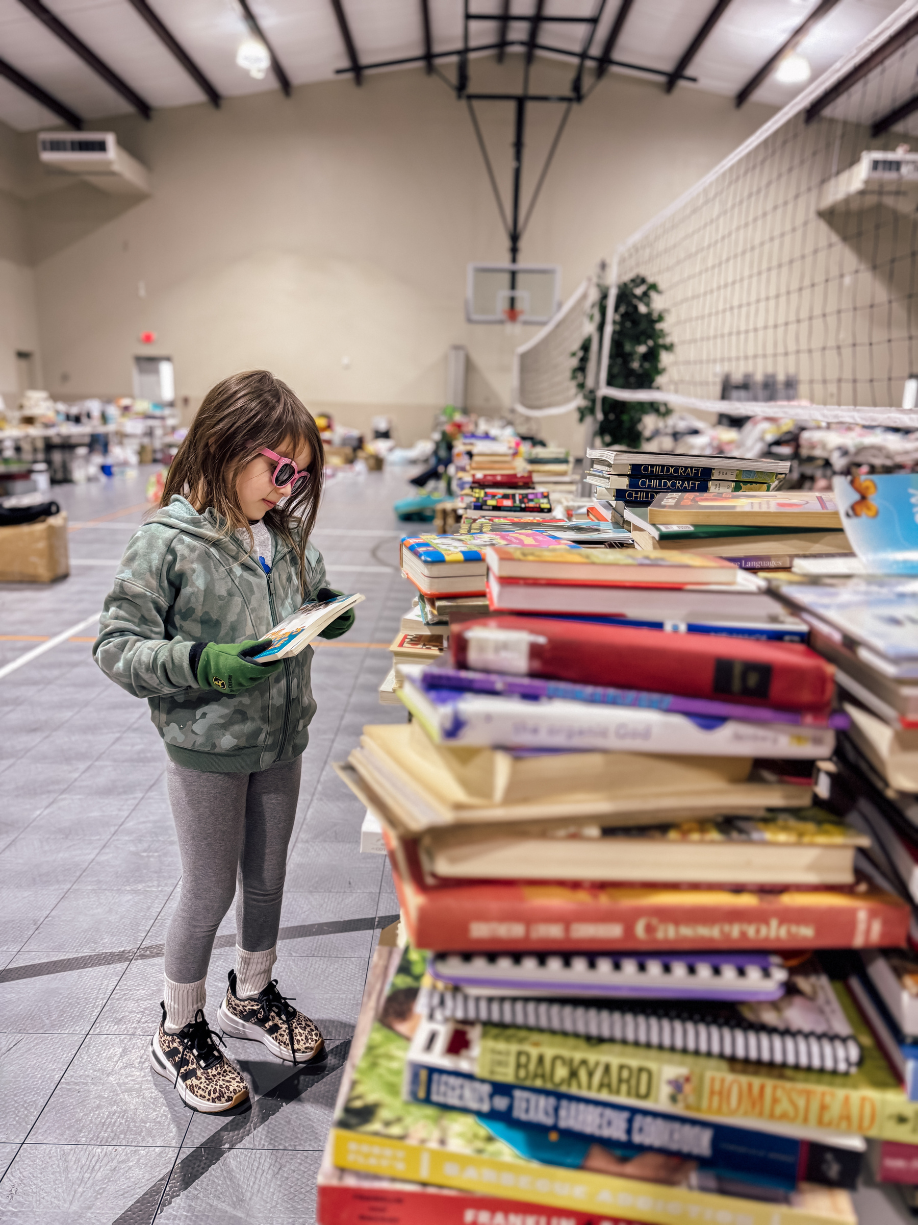 A child in a camouflage jacket and gloves organized a stack of books for garage sale in a gym with a volleyball net.