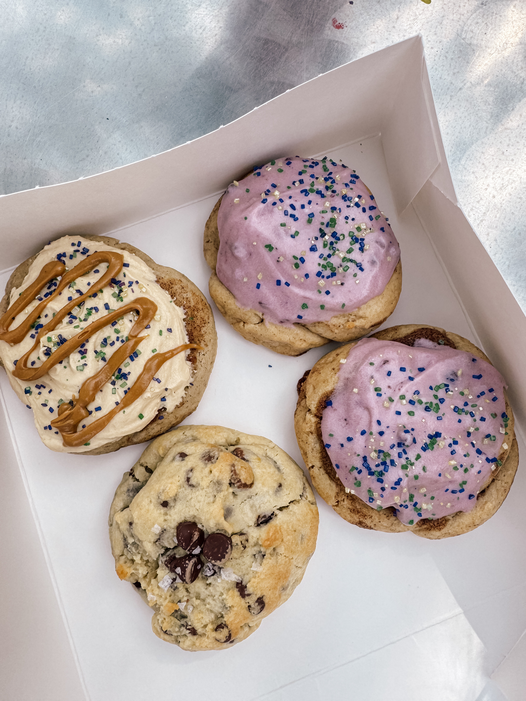 A box contains four assorted cookies, two with purple icing and sprinkles, one with cream icing and caramel drizzle, and one chocolate chip.