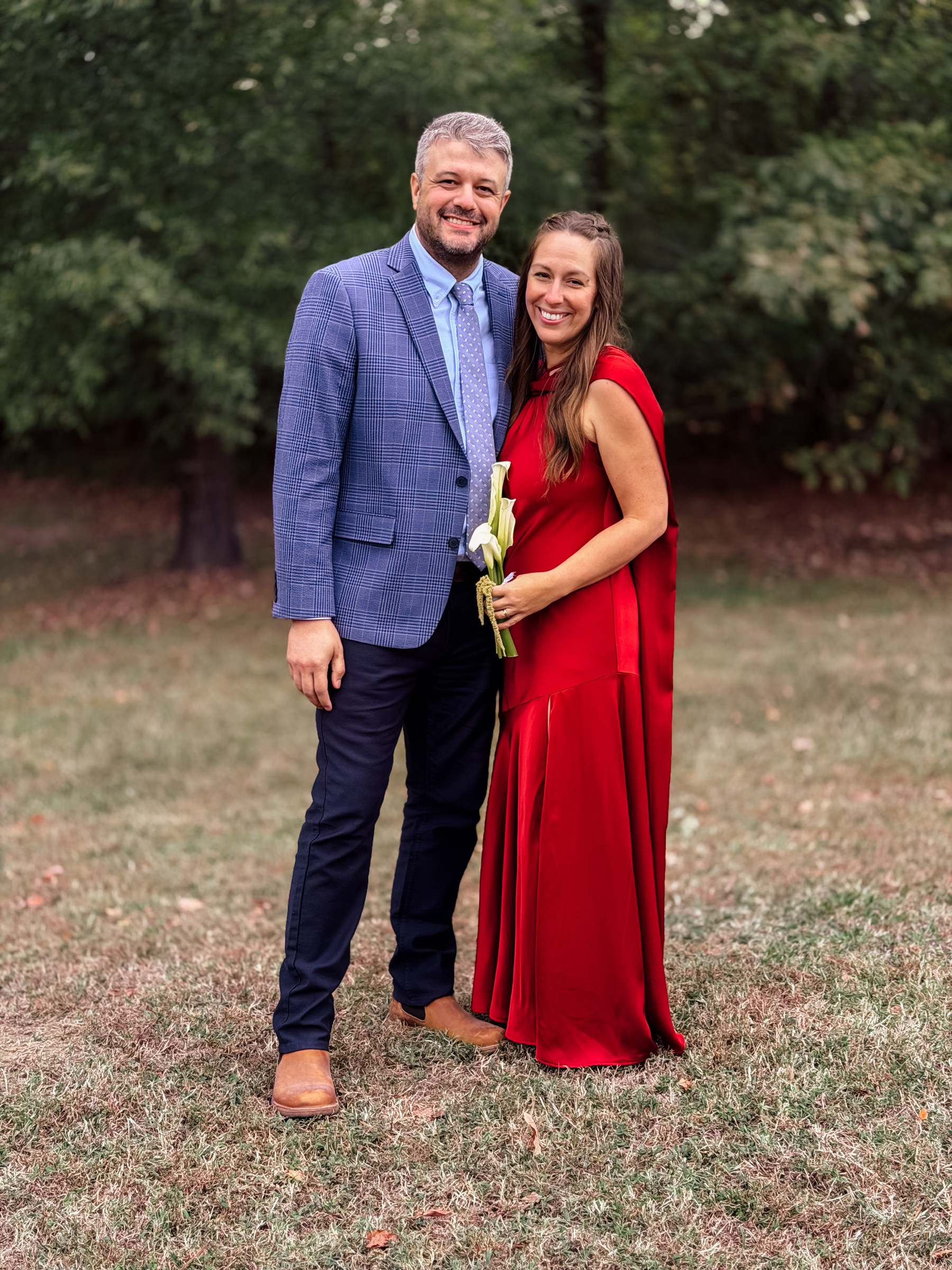 A man in a blue suit and a woman in a red dress stand smiling together on a grassy area with trees in the background.