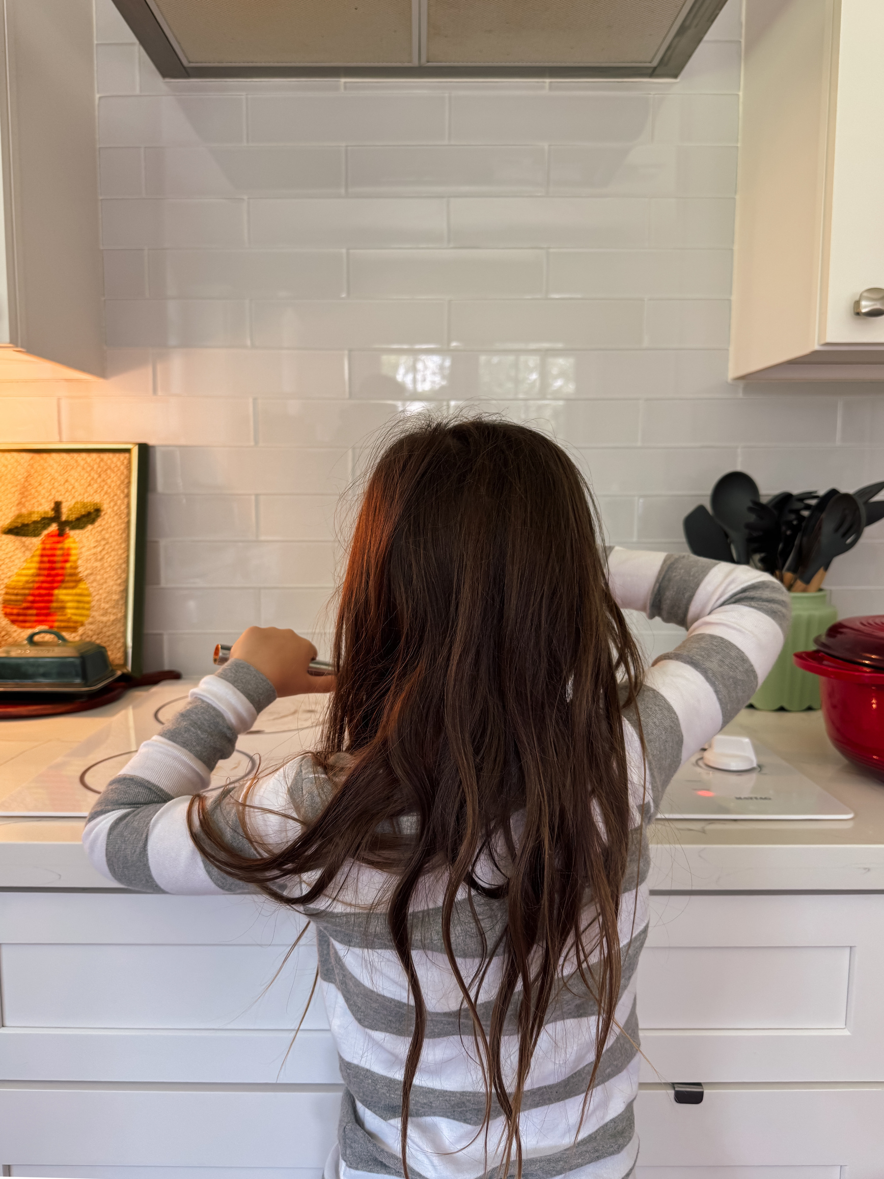 A girl with long hair, wearing a striped pajama top, is standing at a kitchen counter facing the stove, with utensils and a pot nearby.