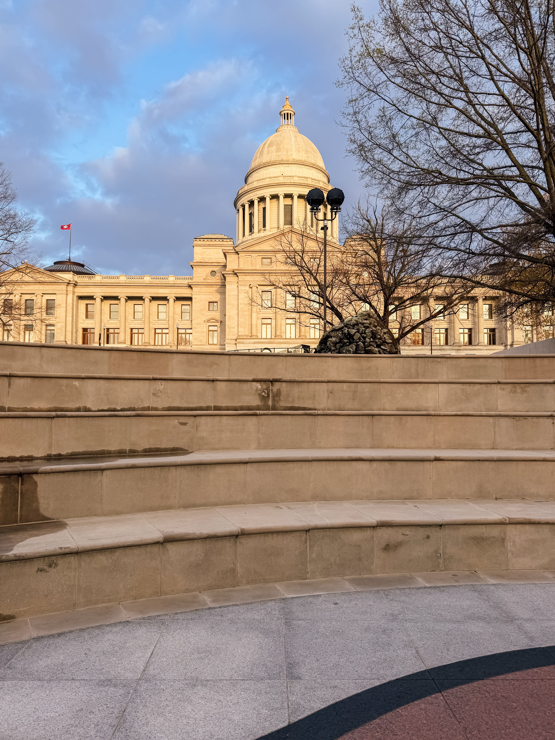 A grand building with a large dome is seen behind a series of stone steps, flanked by bare trees and illuminated by a partly cloudy sky.