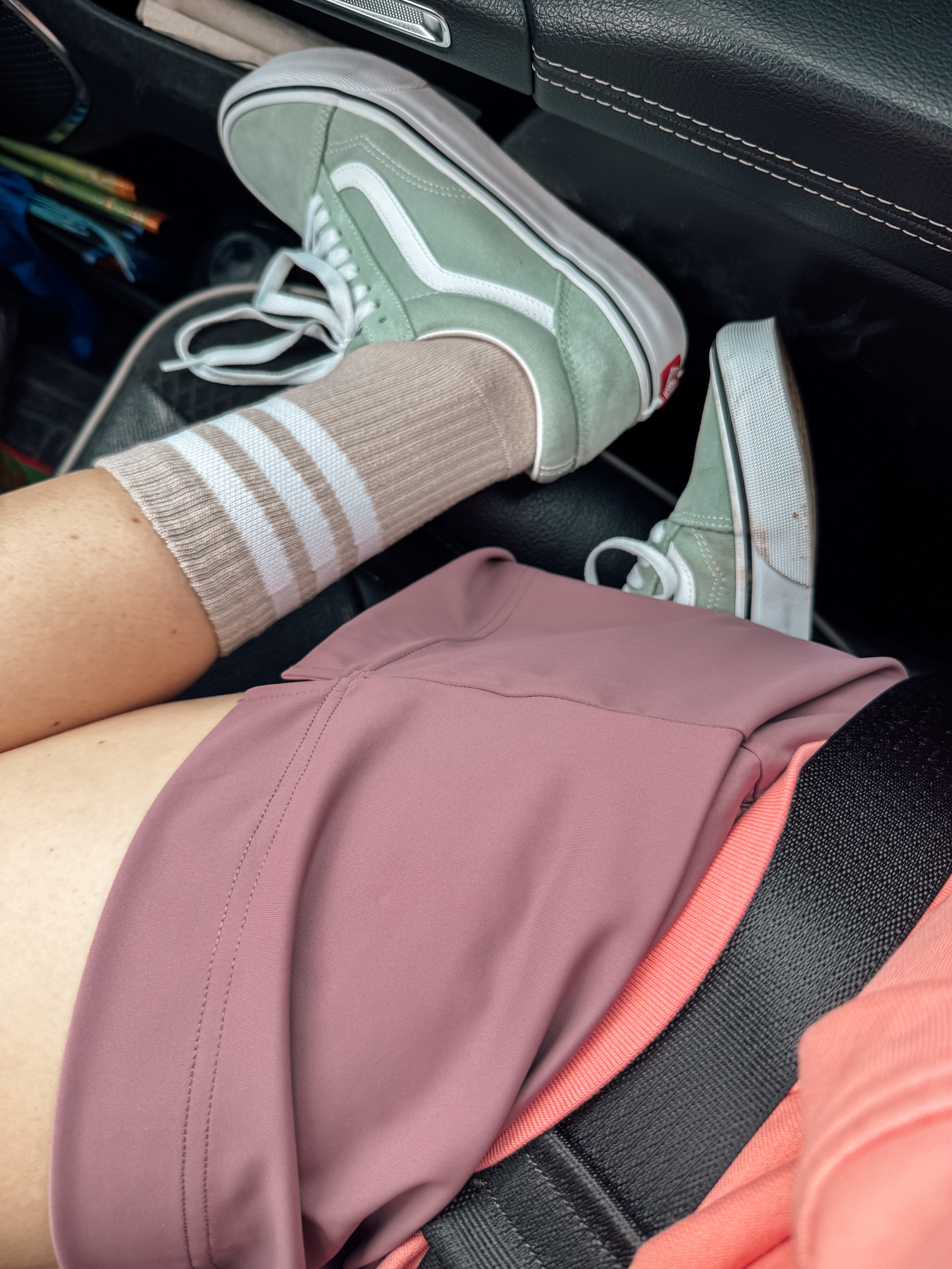 A person wearing green and white sneakers, cream-colored striped socks, and a mauve skirt is seated in a car with their feet pulled up on the seat. 