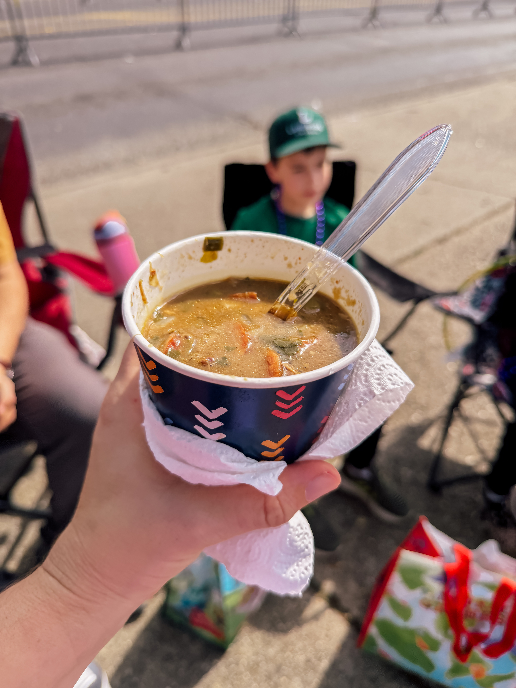 A hand holding a cup of gumbo with a spoon in it, while a person sits blurred in the background.